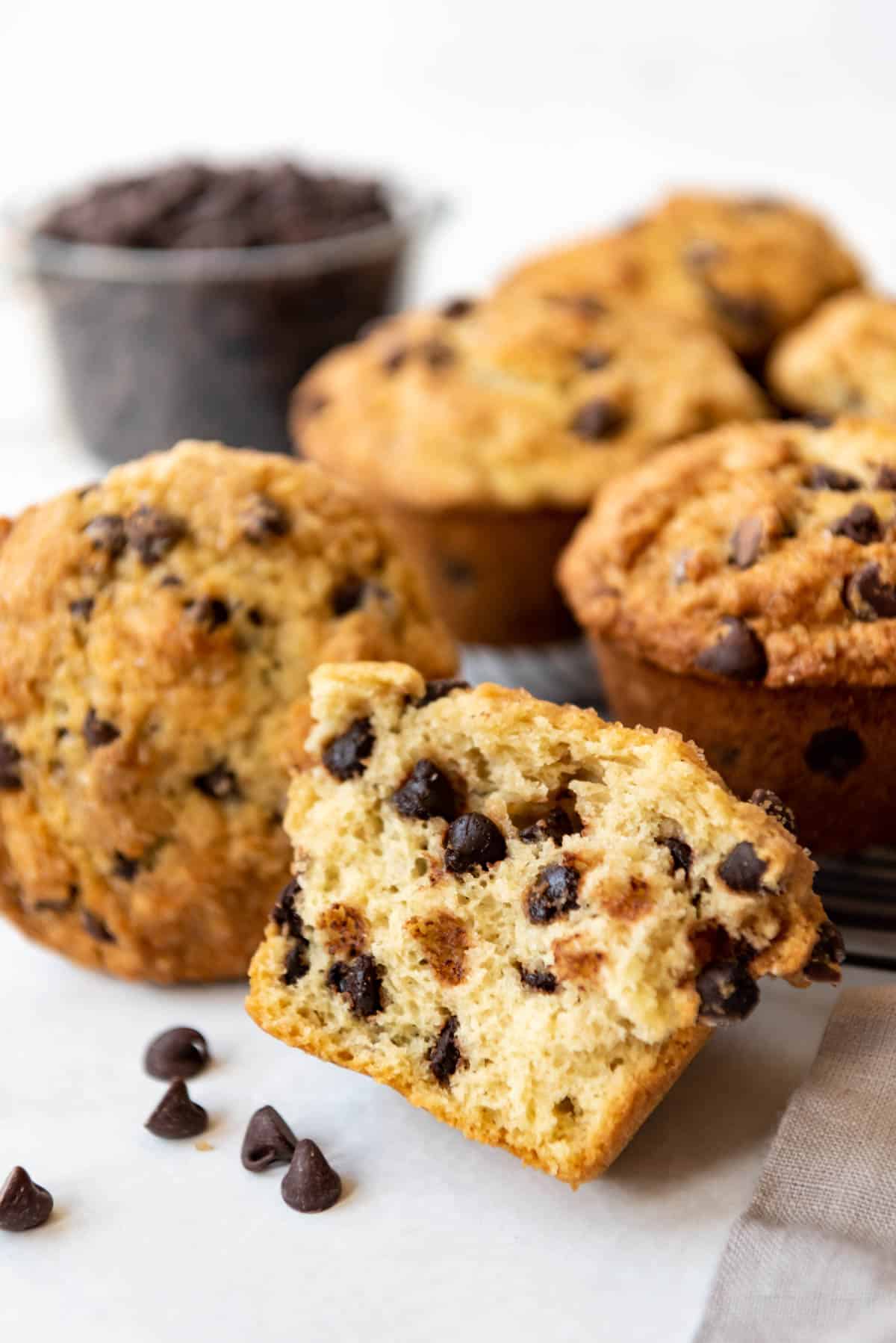 Half of a chocolate chip muffin in front of more muffins and a bowl of chocolate chips.