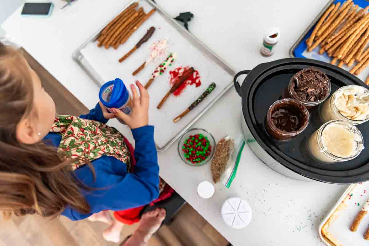 A slow cooker with chocolate in mason jars and girl decorating chocolate covered pretzels.