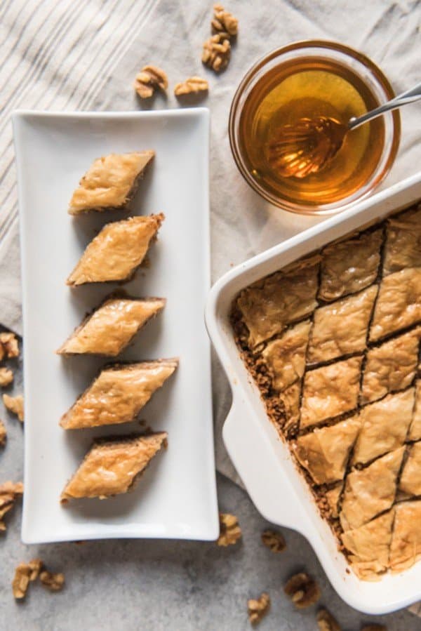 a row of baklava on white plate next to baking dish and bowl of amber colored liquid