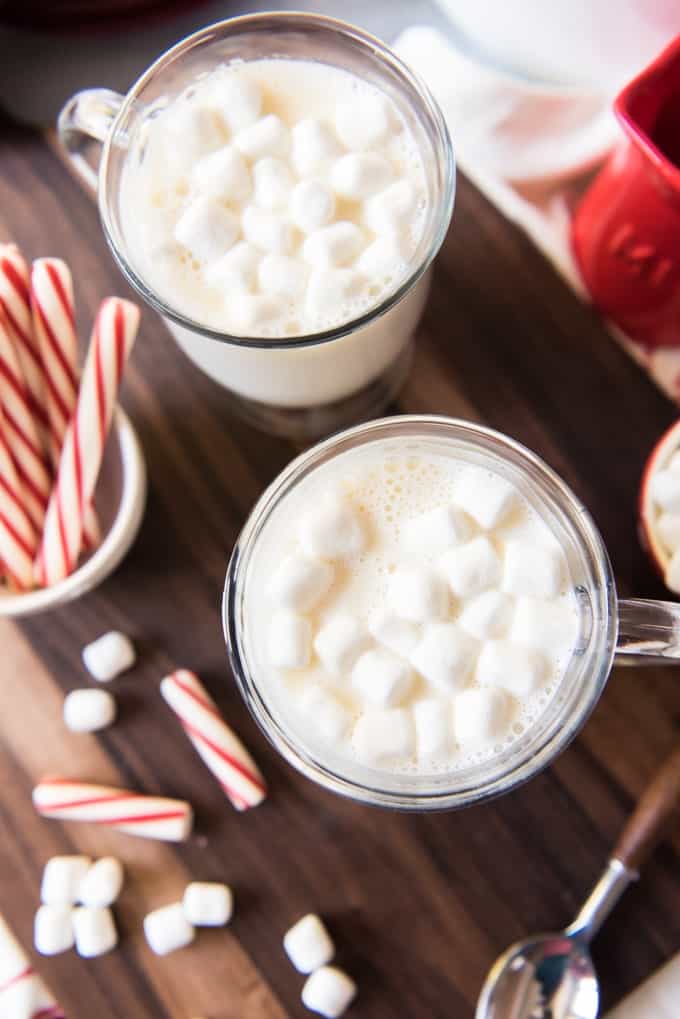 Overhead picture of two mugs of decadent white hot chocolate with marshmallows on top and peppermint sticks for stirring.