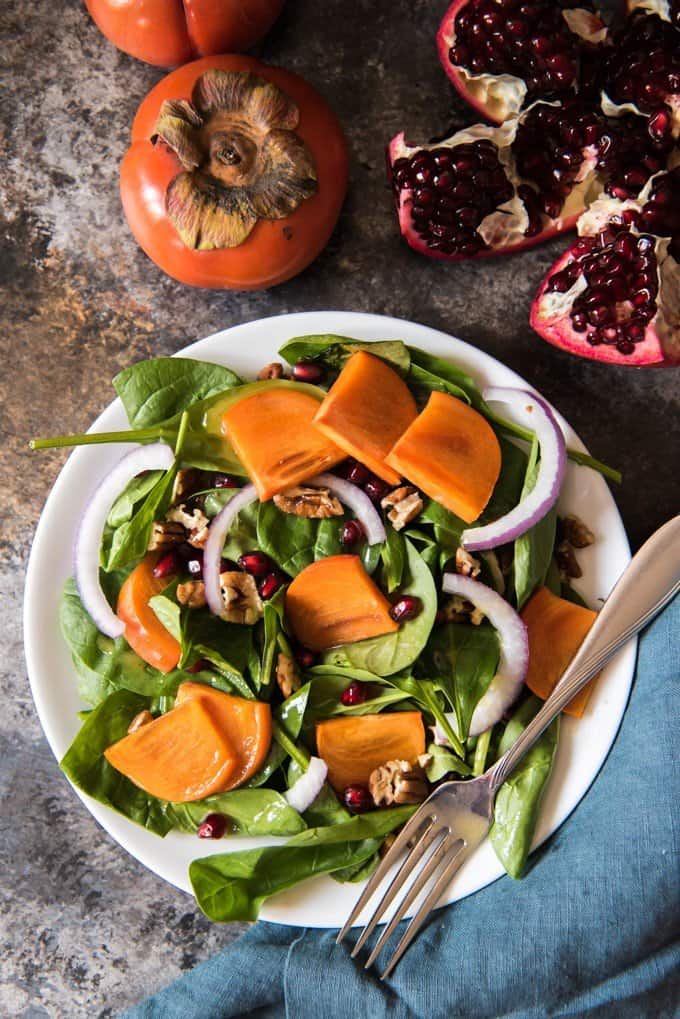 A plate of salad made with sliced persimmons, pomegranate seeds, spinach, red onions, and pecans, with a Maple Citrus Vinaigrette.