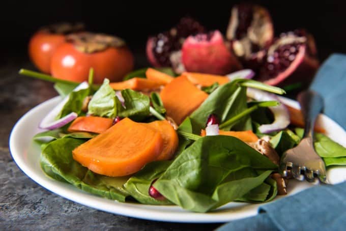 side view of Pomegranate, Spinach, & Persimmon Salad with a fork and fruit in the background