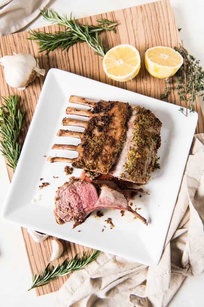 Overhead shot of a square white plate with a roasted rack of lamb on it, crusted in herbs and garlic with two lamb chops sliced off.