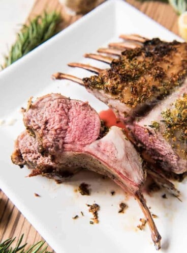 Rosemary & Garlic Oven Roasted Rack of Lamb on a white plate with one chop sliced and turned sideways to reveal meat grain and juiciness