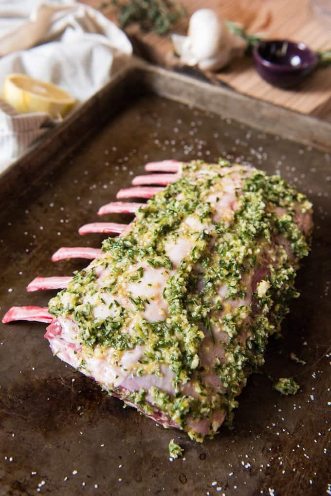 Marinated rack of lamb resting on a baking sheet and ready for roasting in the oven, covered in an olive oil, lemon zest, garlic, rosemary and thyme paste and seasoned with salt and pepper.