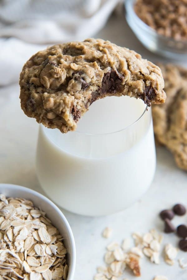 A Toffee Oatmeal Chocolate Chip Cookie with a bite taken out of it, resting on the rim of a glass of milk. 