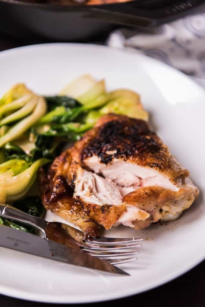 A white plate with a crispy cast iron skillet chicken thigh and stir-fried baby bok choy on it.
