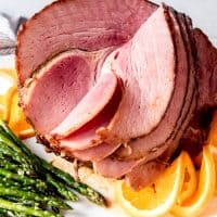 A juicy Brown Sugar Glazed Ham on a white platter with orange slices and asparagus