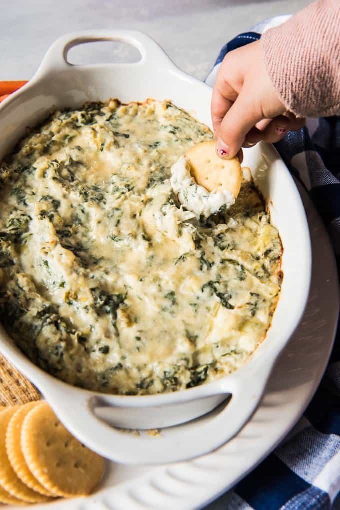 A hand scooping up cheesy spinach artichoke dip with a Ritz cracker.