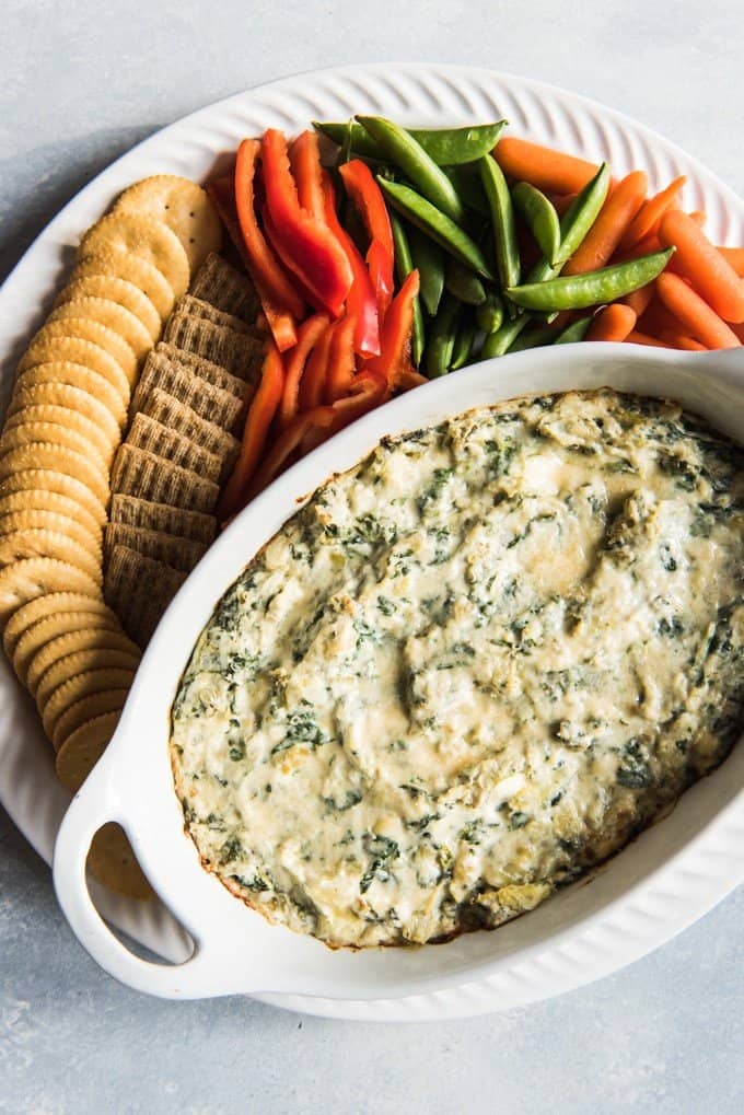 an aerial view of a white baking dish full of spinach artichoke dip with crackers and veggies to the side