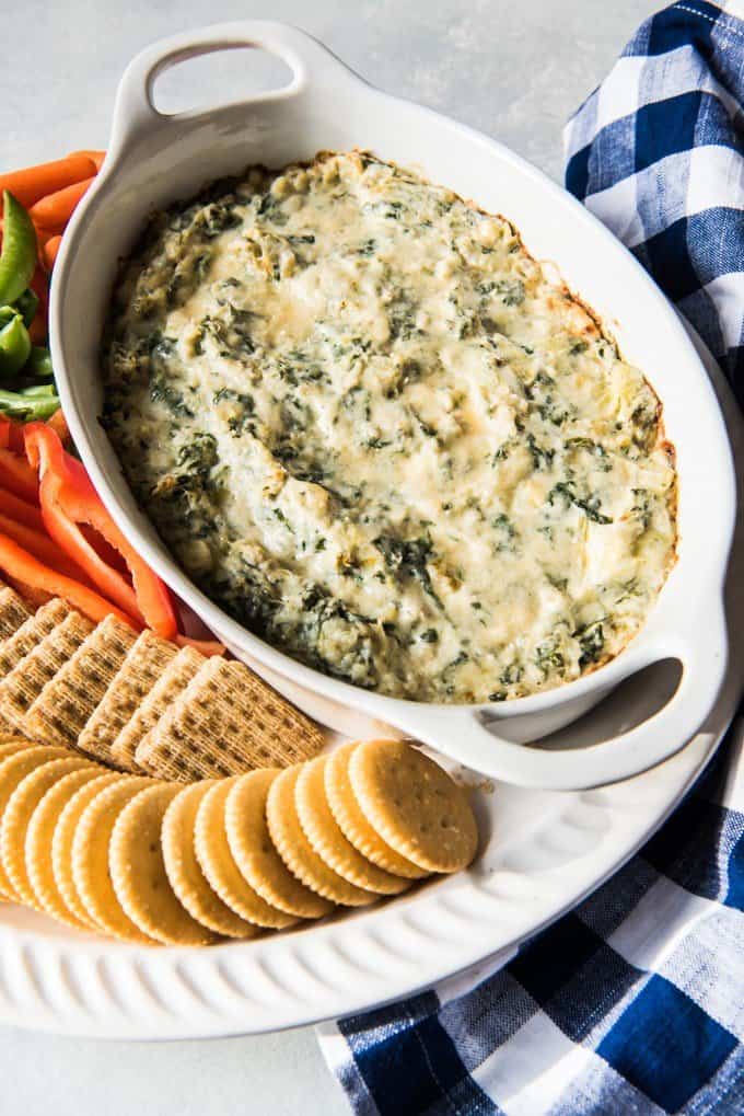 a white baking dish full of spinach dip with crackers and veggies to the side