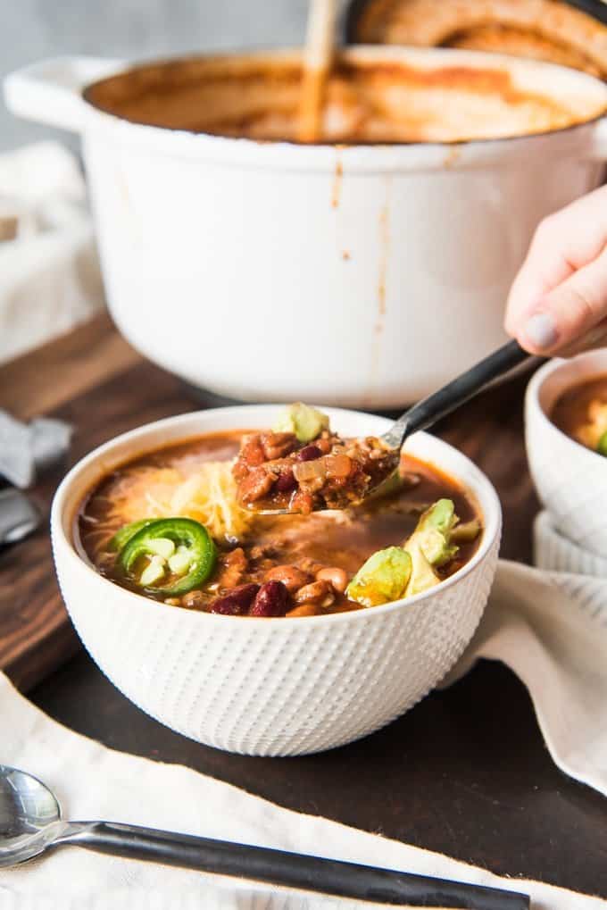 A hand holding a spoonful of chili over a bowl of chili loaded with toppings.