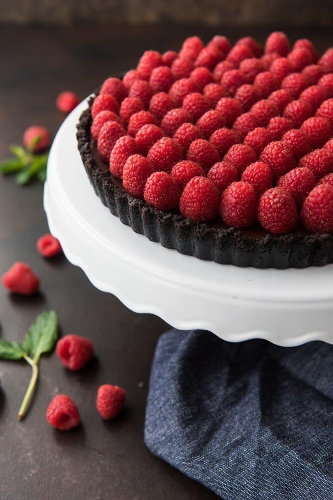 a delicious chocolate and raspberry tart on a white cake stand with scattered berries and leaves below