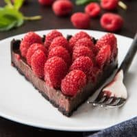 a slice of chocolate raspberry tart on a white plate with a fork and fresh berries and leaves in back