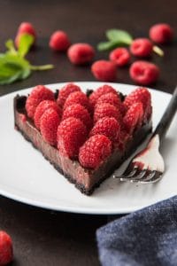 a slice of chocolate raspberry tart on a white plate with a fork and fresh berries and leaves in back