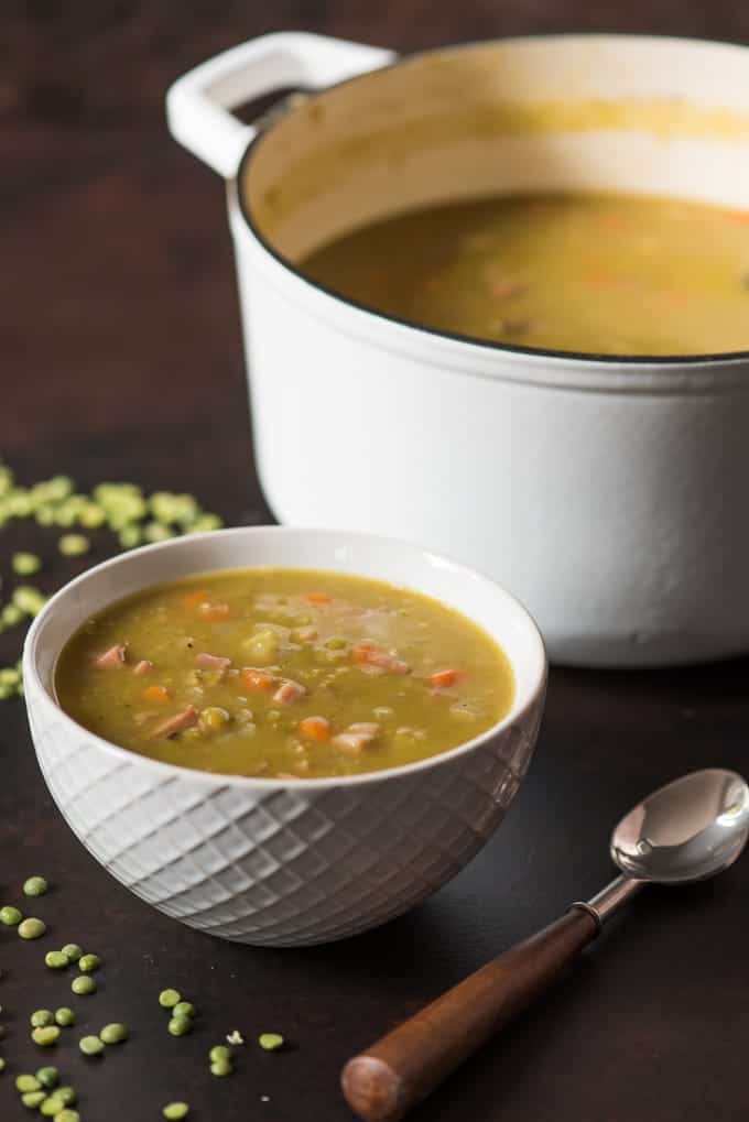 a white bowl of split pea soup next to a pot and spoon with scattered peas around