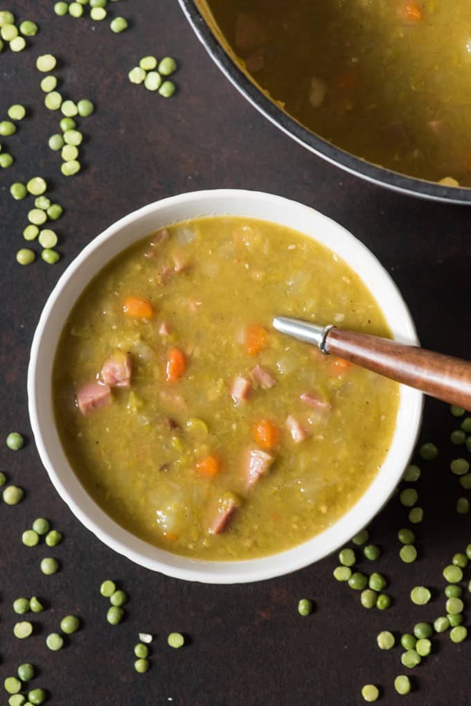A bowl of split pea soup made with ham, surrounded by dried split peas and sitting beside a dutch oven full of soup.