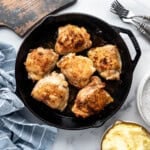 An overhead image of a pan of chicken thighs with a blue cloth napkin, mashed potatoes, a wooden cutting board, and forks nearby.