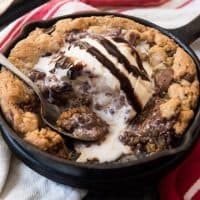 a skillet with a chocolate chip pazookie inside topped with ice cream and chocolate syrup and a spoon removing bites