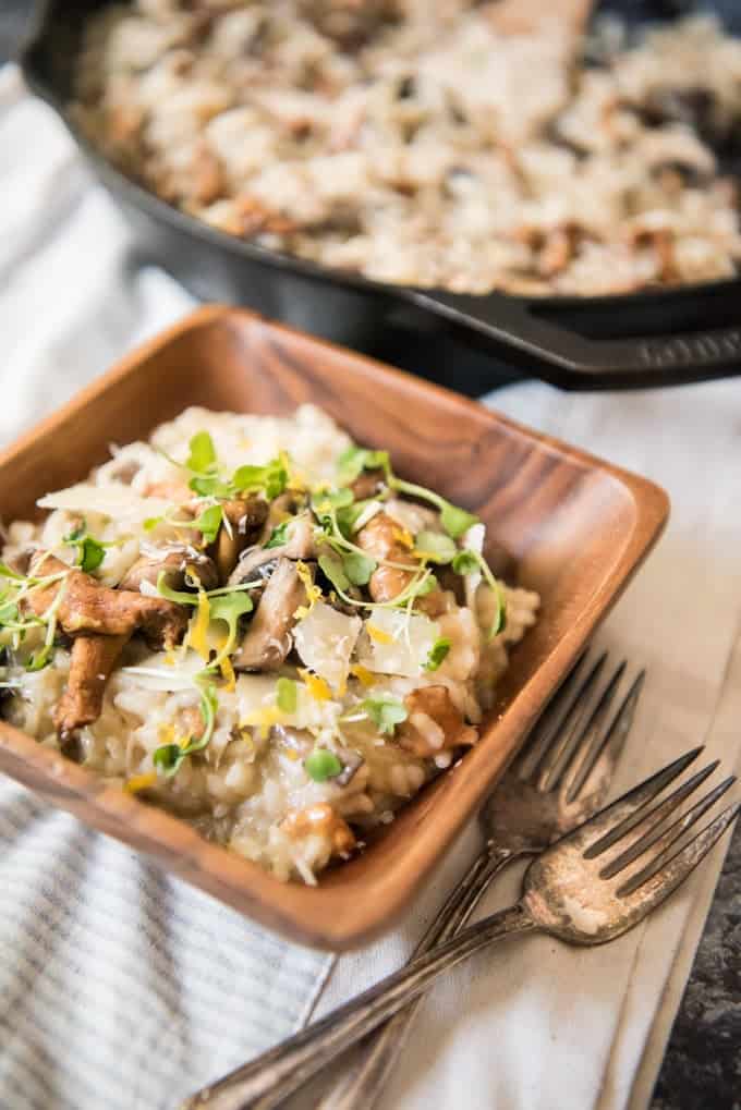 creamy mushroom risotto in a wooden bowl next to two forks and a cast iron pan with more in the background