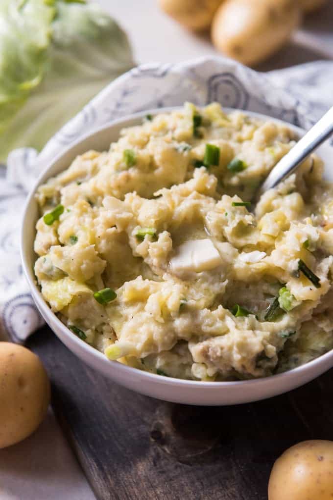 A big bowl of Irish colcannon made with mashed potatoes, sauteed cabbage, and green onions, with butter melting on top and a big serving spoon sticking out of the potatoes.