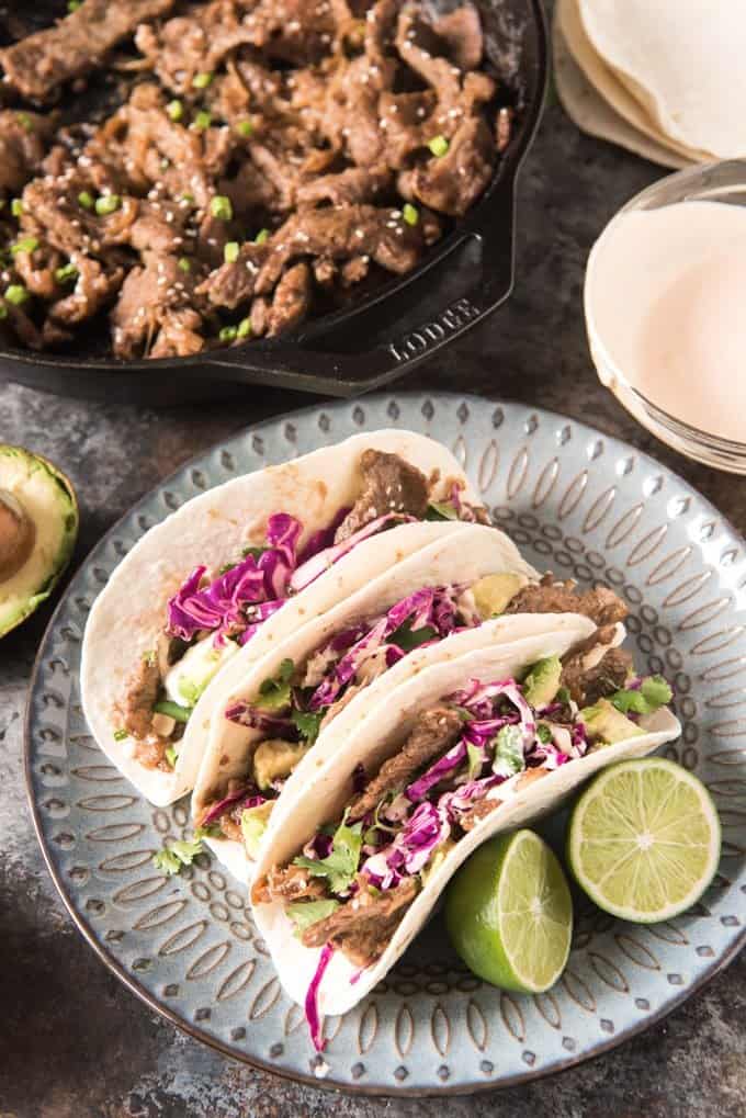 bulgogi tacos on a plate with sliced lime next to a cast iron dish of meat