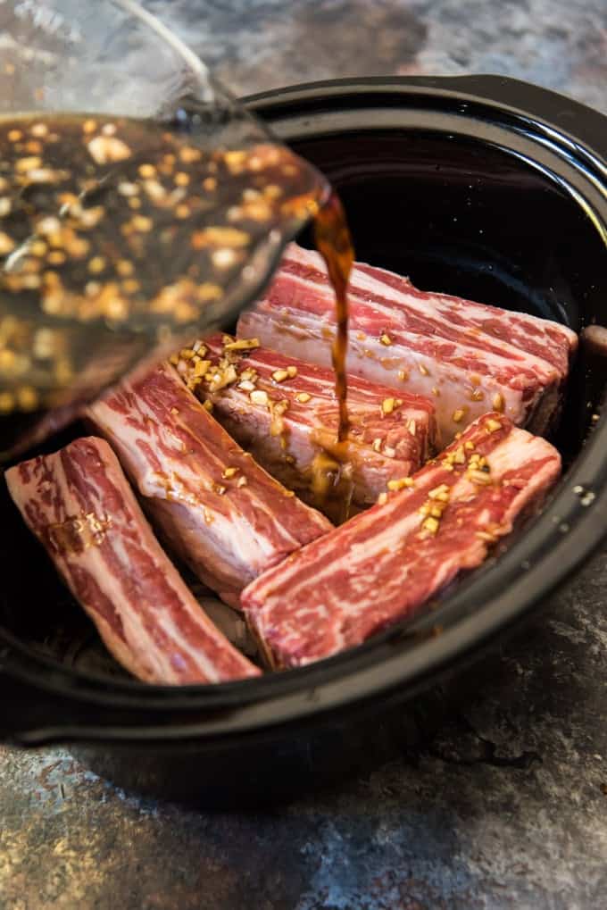Crockpot with beef short ribs and sliced onions in it, with a soy sauce based marinade being poured over the top.