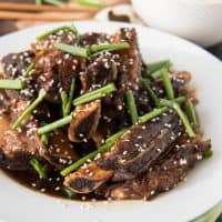 korean beef shortribs topped with sesame seeds and green onion on a white plate with a bowl of rice to the side