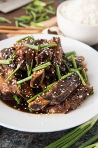 korean beef shortribs topped with sesame seeds and green onion on a white plate with a bowl of rice to the side