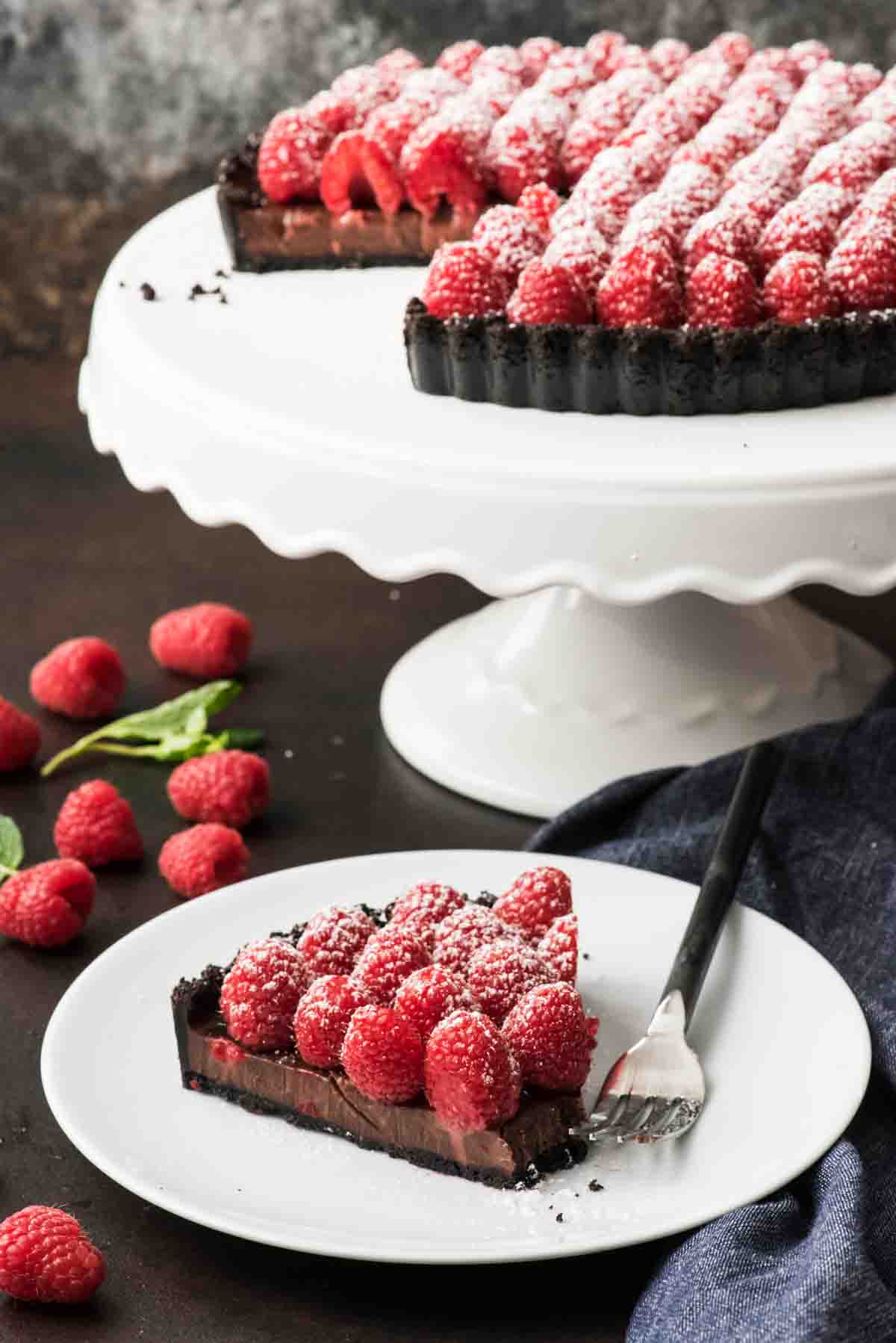 A slice of raspberry chocolate tart on a plate in front of the rest of the tart on a white cake stand.