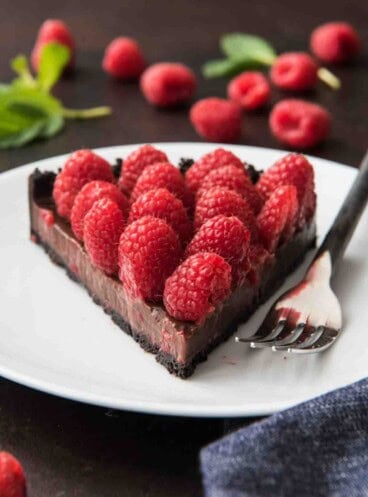 A slice of raspberry chocolate tart with an oreo cookie crust, chocolate ganache and fresh raspberries on a white plate with a fork.