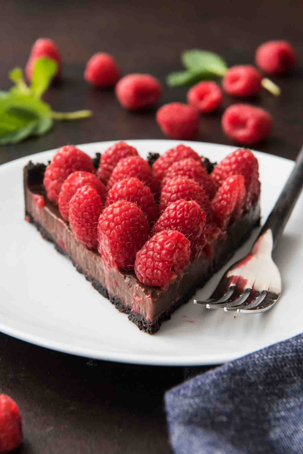 A slice of raspberry chocolate tart with an oreo cookie crust, chocolate ganache and fresh raspberries on a white plate with a fork.