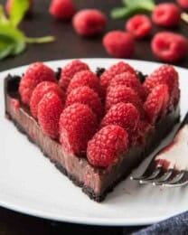 A slice of raspberry chocolate tart with an oreo cookie crust, chocolate ganache and fresh raspberries on a white plate with a fork.