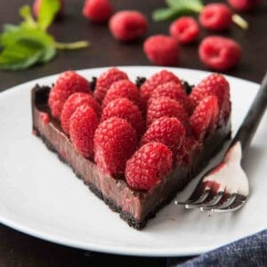 A slice of raspberry chocolate tart with an oreo cookie crust, chocolate ganache and fresh raspberries on a white plate with a fork.