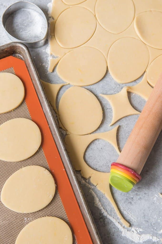 An image of cut-out sugar cookies on a silpat mat, next to rolled sugar cookie dough cut into easter egg shapes.