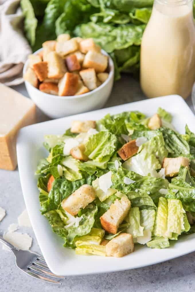 caesar salad on a white plate in front of a bowl of crutons and bottle of dressing