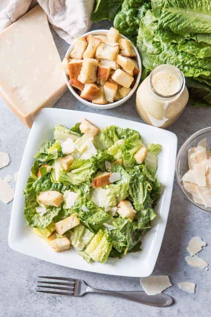 An image of a original caesar salad on a white plate with extra croutons and caesar salad dressing next to it.