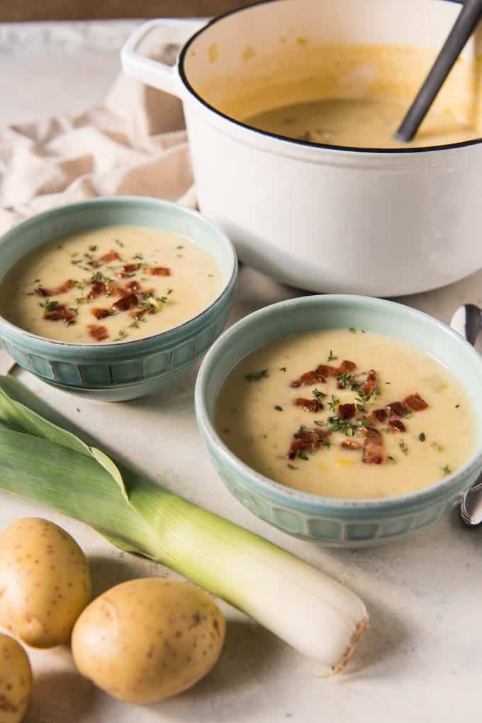 An image of a white pot of potato leek soup with two bowlfuls of soup and a leek and potatoes off to the side.