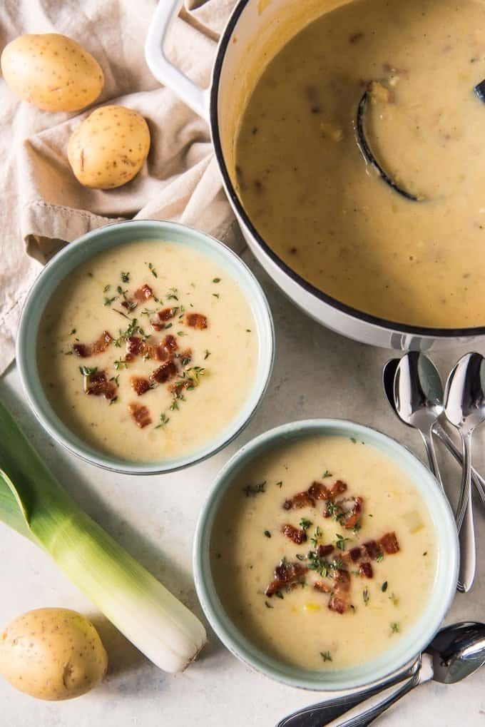 Two bowls of creamy Irish leek and potato soup garnished with crispy bacon and thyme leaves, next to a pot of leek soup.
