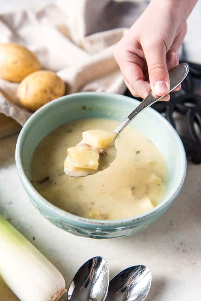 An image of a hand holding up a spoonful of creamy Irish leek and potato soup with chunks of potato in it over a bowlful of the soup.