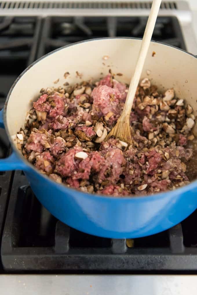 An image of a dutch oven on a stove filled with ground beef, ground pork, pancetta, and mushrooms for a bolognese sauce recipe.