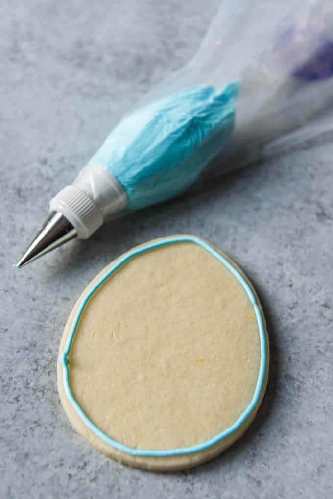 An image of an egg shaped sugar cookie with a border of blue royal icing piped around the edge.