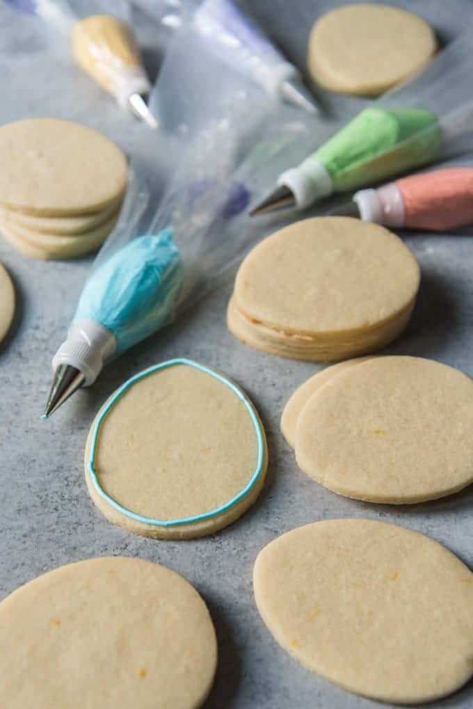 An image of egg-shaped sugar cookies spread out for decorating with royal icing, with one cookie having a border of blue royal icing already piped around the edges.
