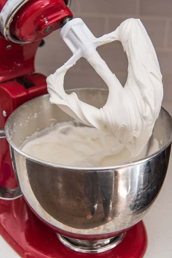 An image of white royal icing in a stand mixer with the beater lifted out of the mixing bowl to show how thick the icing is.