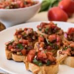 Small rounds of toasted baguette arranged on a white plate and topped with homemade Italian bruschetta.