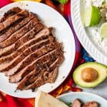 sliced carne asada on a white plate next to a halved avocado, tortillas, and more