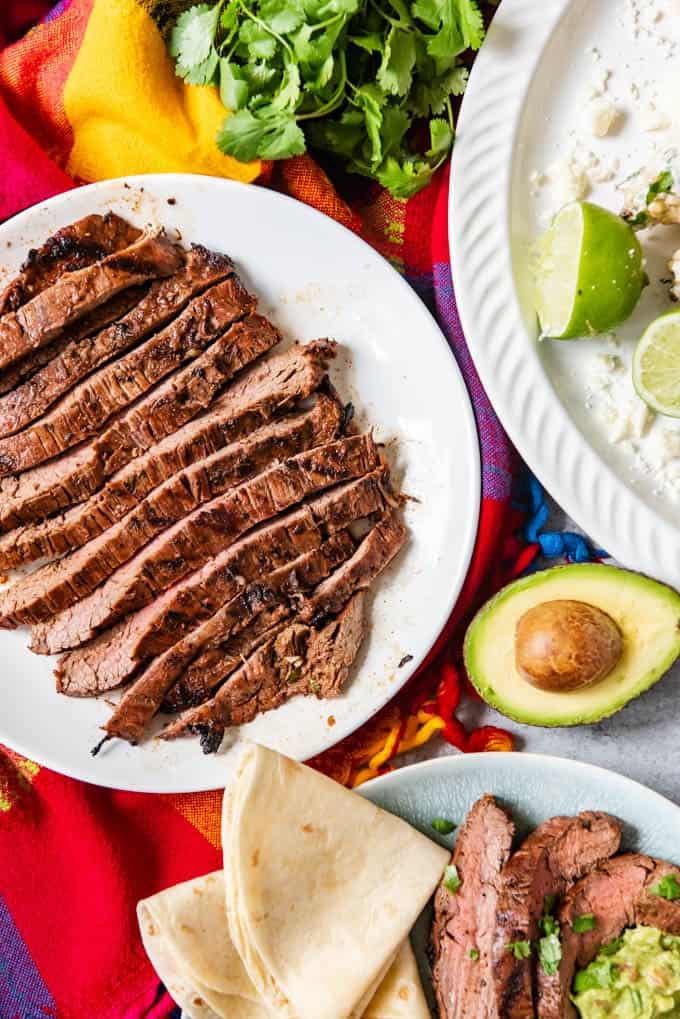 sliced carne asada on a white plate next to a halved avocado, tortillas, and more