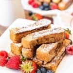 a pile of french toast sticks on a white plate topped with powdered sugar and garnished with fresh berries