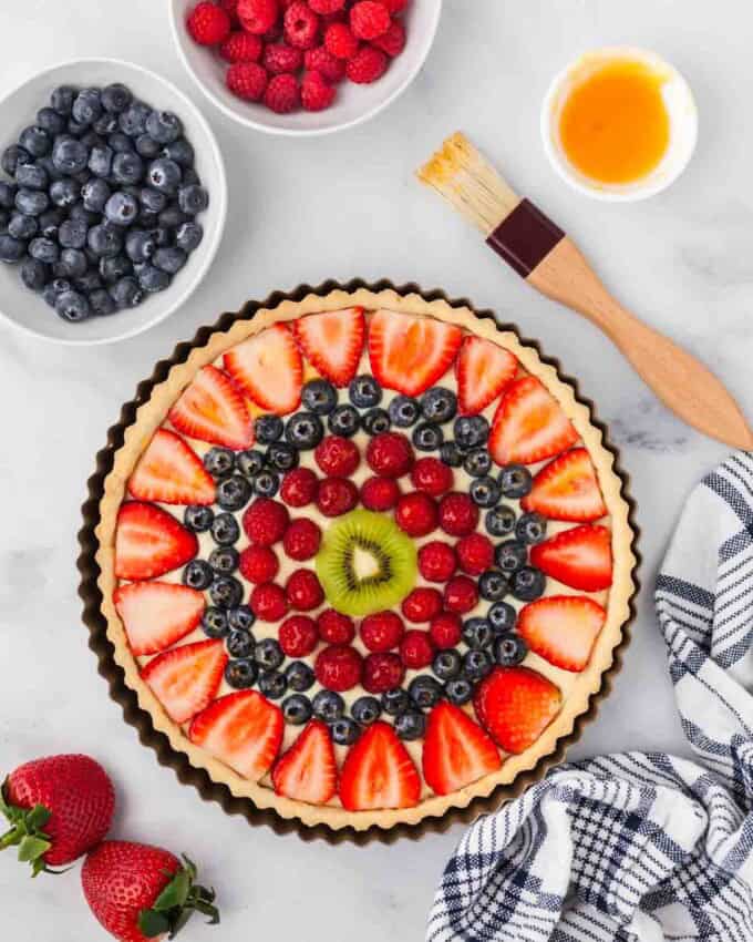 A finished fruit tart next to bowls of fruit and a pastry brush.