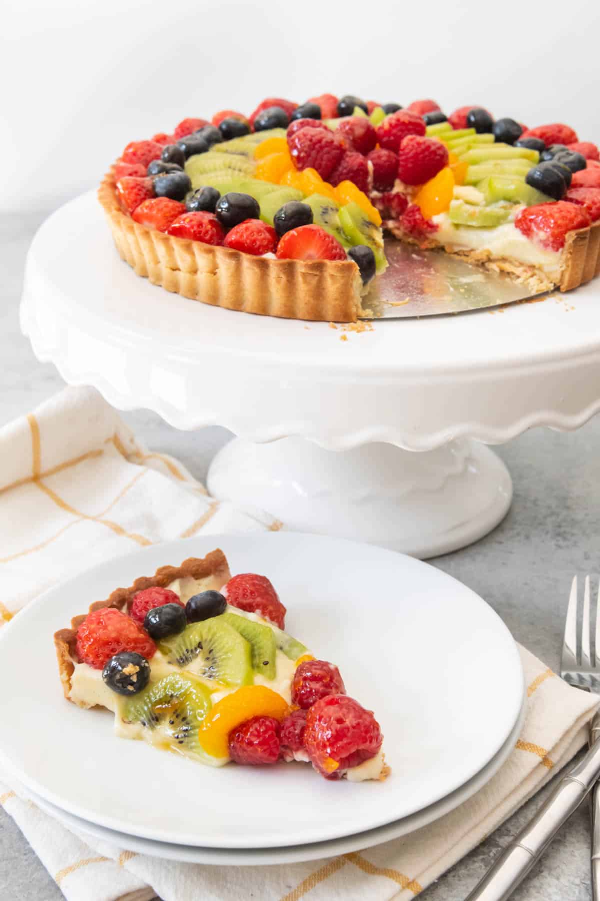 An image of a fresh fruit tart on a white cake stand, with one slice cut and served on a white plate. 